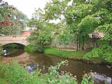 A river flowing under a stone bridge with trees and plants along the riverbank at Dunlin Cottage in Bamburgh