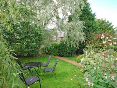 A garden with metal chairs and a table on grass surrounded by trees and bushes at Dunlin Cottage in Bamburgh