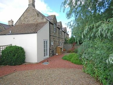 A stone cottage with a white extension and a garden path surrounded by bushes and trees at Dunlin Cottage in Bamburgh