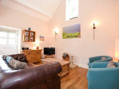 A living room with a brown leather sofa two blue armchairs a wooden coffee table a wooden cabinet with shelves a small television and wall art at High Tide in Newton-by-the-Sea