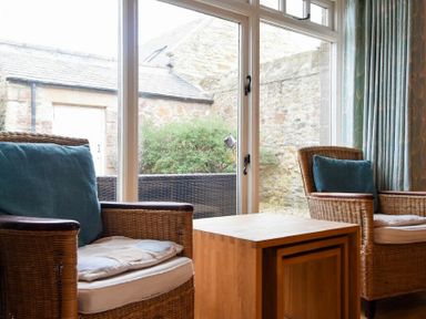 A sitting area with two wicker armchairs with cushions and a wooden nesting table in front of large windows at High Tide in Newton-by-the-Sea