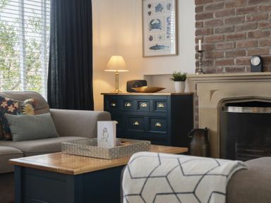 A living room with a sofa a wooden coffee table a dark dresser with a lamp and a fireplace at Sea Croft in Beadnell