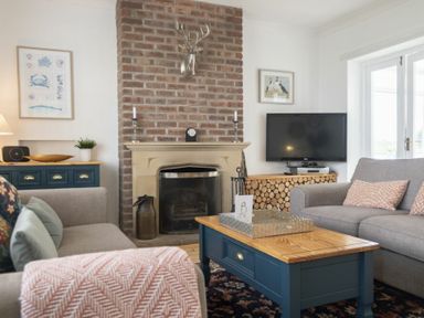 A living room with a brick fireplace a blue coffee table two grey sofas and a television at Sea Croft in Beadnell