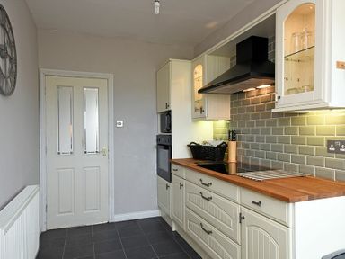 A kitchen with white cabinets wooden countertop electric stove gray tiled backsplash and a door at Sea Croft in Beadnell