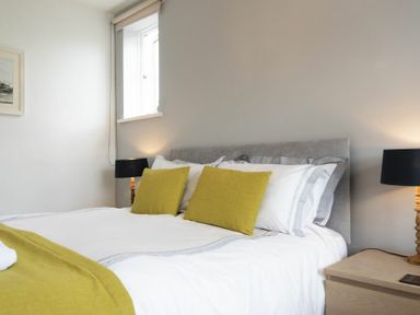 A bedroom with a bed with white linens and yellow cushions and a bedside table with a lamp at Sea Croft in Beadnell