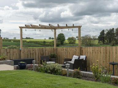 An outdoor garden seating area with wicker chairs and cushions under a wooden pergola with hanging lights at Sea Croft in Beadnell