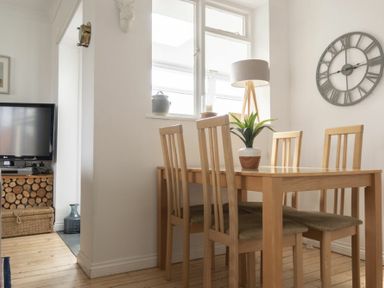 A dining area with a wooden table and four chairs a plant a floor lamp and a wall clock at Sea Croft in Beadnell