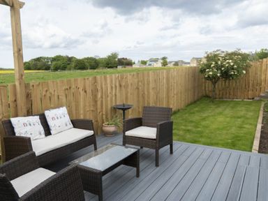 A backyard with wicker patio furniture on a gray deck and a wooden fence surrounding a grassy area with a small tree at Sea Croft in Beadnell