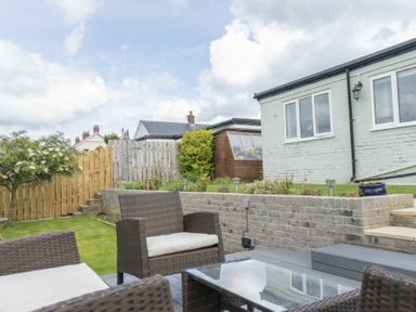An outdoor patio with wicker chairs and a glass table next to a brick retaining wall and a garden shed at Sea Croft in Beadnell