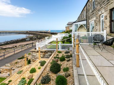 A stone building with a patio and garden overlooking a pier and the sea at Old Coastguard Cottages in Amble
