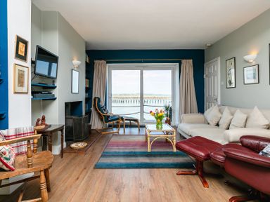 A living room with beige sofa red leather chair coffee table and sliding glass door overlooking water at Old Coastguard Cottages in Amble