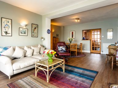 A living room with a beige sofa, a glass coffee table, a colorful rug, a red armchair, a wooden dining table with chairs, and wall art at Old Coastguard Cottages in Amble