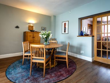 A dining room with a round wooden table and six chairs on a round rug at Old Coastguard Cottages in Amble