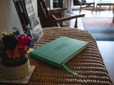 A green guest book on a wicker table next to a vase with colorful flowers in a living room at Old Coastguard Cottages in Amble