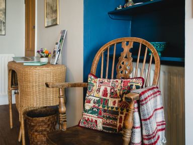 A wooden chair with a patterned cushion and striped blanket next to a wicker table with books and flowers at Old Coastguard Cottages in Amble