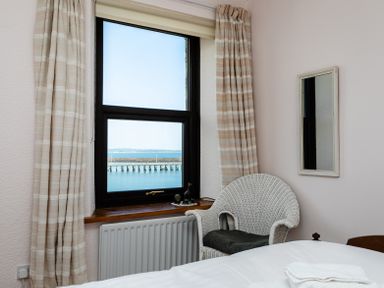 A bedroom with a window showing a view of water and a pier a white wicker chair with a cushion beige curtains and a wall mirror at Old Coastguard Cottages Amble