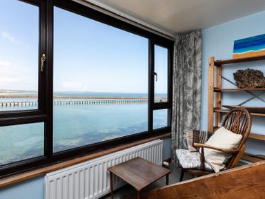 A room with a wooden chair and small table next to a large window overlooking water at Old Coastguard Cottages in Amble
