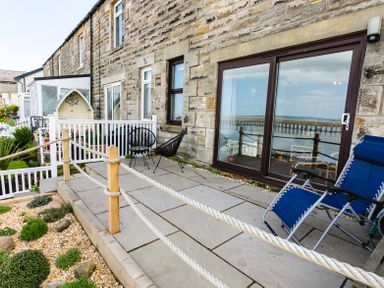 An outdoor patio with stone flooring two black chairs and one blue lounge chair outside a stone house with large glass sliding doors showing water view at Old Coastguard Cottages in Amble