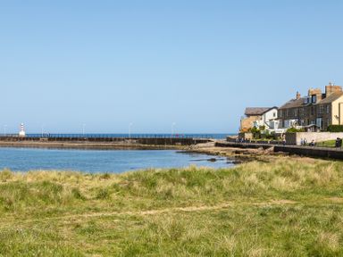 A coastal scene with grassy foreground a body of water a stone pier with a lighthouse and a row of houses at Old Coastguard Cottages in Amble