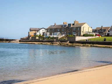 A sandy beach with calm water and a row of stone and brick houses along the shore at Old Coastguard Cottages in Amble