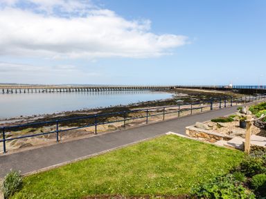 A paved walkway with railings along a rocky shoreline and a long wooden pier at Old Coastguard Cottages in Amble