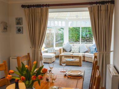 A dining area with wooden chairs and table set with plates and glasses looking into a sitting area with wicker furniture and cushions at Armstrong Cottages No30 Bamburgh