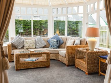 A conservatory with wicker furniture a beige sofa with cushions a wicker coffee table and a side table with a lamp at Armstrong Cottages No30 in Bamburgh