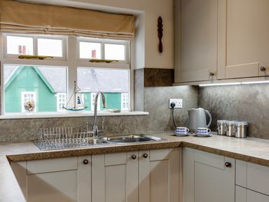 A kitchen sink with a drying rack and a faucet below a window with blinds next to a counter with a kettle cups and canisters at Armstrong Cottages No30 Bamburgh
