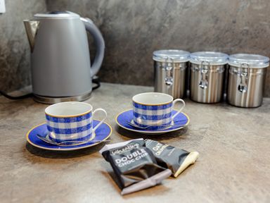A kitchen counter with two blue and white checkered teacups and saucers with spoons a grey electric kettle three metal canisters and two snack bars at Armstrong Cottages No30 Bamburgh