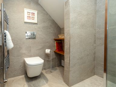 A bathroom with a wall-mounted toilet a towel rack with towels a toilet paper holder and wooden shelves with books at Armstrong Cottages No30 in Bamburgh