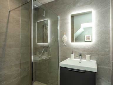 A bathroom with a glass shower enclosure a rectangular illuminated mirror and a white sink at Armstrong Cottages No30 Bamburgh