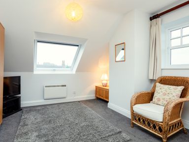 A room with a wicker chair and a floral cushion a TV on a stand a wooden chest with a table lamp a slanted window and a wall heater at Armstrong Cottages No30 in Bamburgh