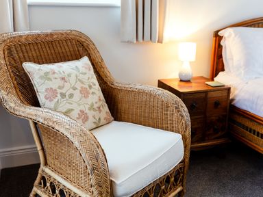 A wicker armchair with a floral cushion next to a wooden bedside table with a lamp and a bed at Armstrong Cottages No30 in Bamburgh