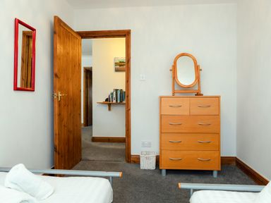 A bedroom with two beds, a wooden dresser with a mirror, an open wooden door, and a shelf with books at Armstrong Cottages No30 in Bamburgh
