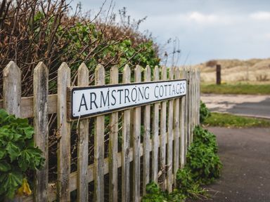 A wooden picket fence with a sign that reads Armstrong Cottages next to bushes and a road at Armstrong Cottages No30 Bamburgh