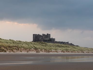 A castle behind sand dunes and a beach under a cloudy sky at Armstrong Cottages No30 in Bamburgh