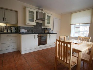 A kitchen with white cabinets black countertop and wooden dining table with chairs near window at Lobster Pot Cottage in Seahouses