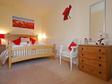 A bedroom with a wooden bed with red cushions a white chair with a red pillow a wooden chest of drawers a mirror and teddy bear at Lobster Pot Cottage in Seahouses