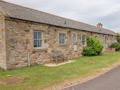 A stone house with multiple windows a wooden bench and bushes on a grassy lawn at Beachward in Embleton