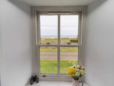 A window with a view of grass road and ocean with binoculars and a vase of flowers on the windowsill at Beachward in Embleton