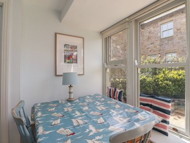 A dining area with a table covered by a blue tablecloth with sailboat patterns a lamp on the table striped cushions on window seats and a framed picture on the wall at Beachward in Embleton