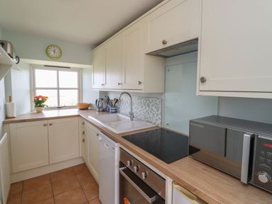 A kitchen with white cabinets a microwave an oven a stovetop a sink a window with a clock and a flower pot on the counter at Beachward in Embleton