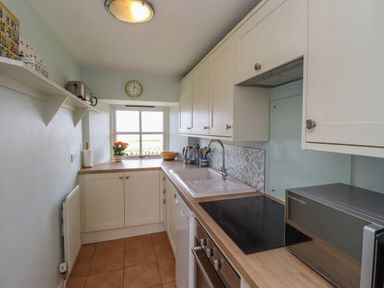 A galley kitchen with white cabinets a sink an electric stovetop a microwave a toaster a kettle and a window with a clock above at Beachward in Embleton
