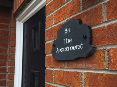 A black door with white frame and a black sign with white text on a brick wall at The Apartment in Bamburgh