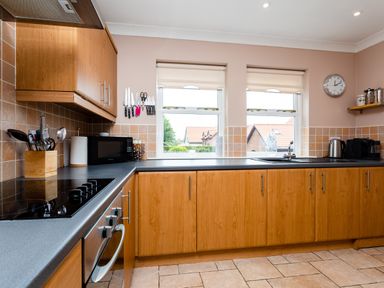 A kitchen with wooden cabinets a black countertop two windows a clock and various kitchen appliances at The Apartment in Bamburgh