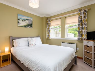 A bedroom with a double bed bedside table lamp windows with curtains and a small chest of drawers at The Apartment in Bamburgh