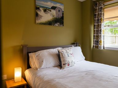 A bedroom with a bed white bedding nightstand with lamp painting on wall window with patterned curtains at The Apartment in Bamburgh
