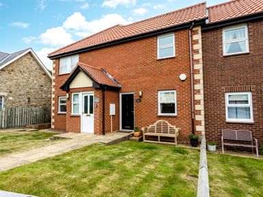 A front view of a brick house with a tiled roof a pathway leading to two doors and benches on grass at The Apartment in Bamburgh