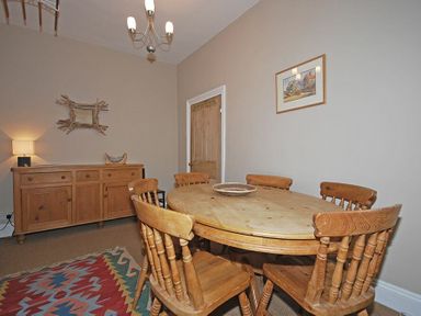 A dining room with a round wooden table and six wooden chairs a wooden sideboard a colorful rug a framed picture and a wall mirror at Beach House (Alnmouth) in Alnmouth