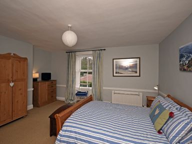 A bedroom with a wooden bed, striped bedding, wooden wardrobe, chest of drawers, window with curtains, and framed pictures on walls at Beach House (Alnmouth) in Alnmouth
